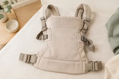 Porte-bébé ventral tissé beige posé seul dans un décor lumineux aux matières naturelles.