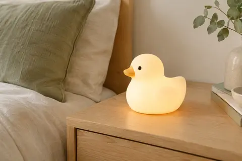 Veilleuse en silicone souple en forme de canard, allumée sur une table de chevet en bois clair.
