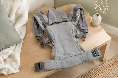 Porte-bébé évolutif en coton gris posé à plat dans une chambre lumineuse aux tons doux.
