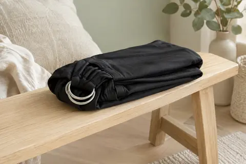 Écharpe de portage Momcozy noire avec anneaux, pliée sur un banc en bois clair