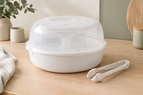Stérilisateur de biberons blanc pour micro-ondes avec sa pince, posé sur un plan de travail clair.