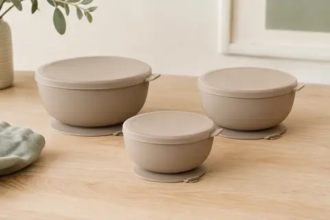 Trois bols bébé en silicone brun clair avec couvercles et bases à ventouse, posés sur une table claire.